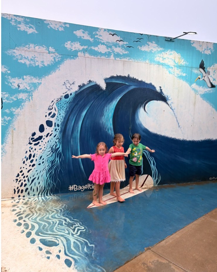 Three young children standing on a painted surfboard in front of a large wave mural on an exterior wall