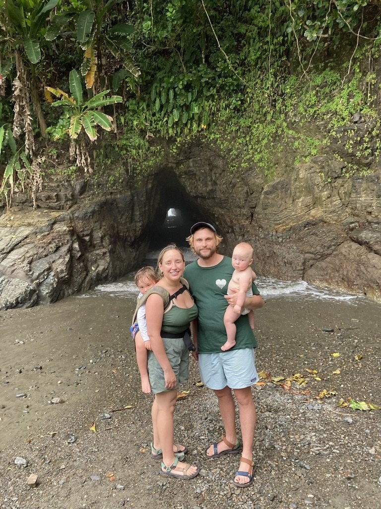 A family standing at the mouth of the sea cave at Playa Ventanas in July 2023, with a three-year-old in a back carrier and a one-year-old held in his father's arms