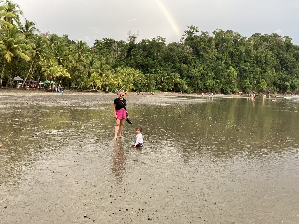 Lela standing beside her one-year-old granddaughter on the low-tide flats at Playa Ventanas in July 2023, a rainbow over the jungle headland behind them and vendor tents along the treeline