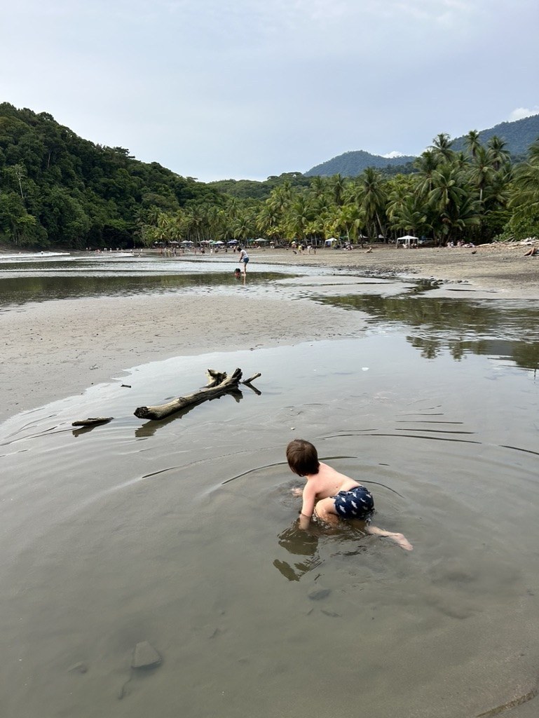 A three-year-old sitting in the shallow river pool at Playa Ventanas in July 2023, with a driftwood log beside him and the low-tide flats and vendor umbrellas behind