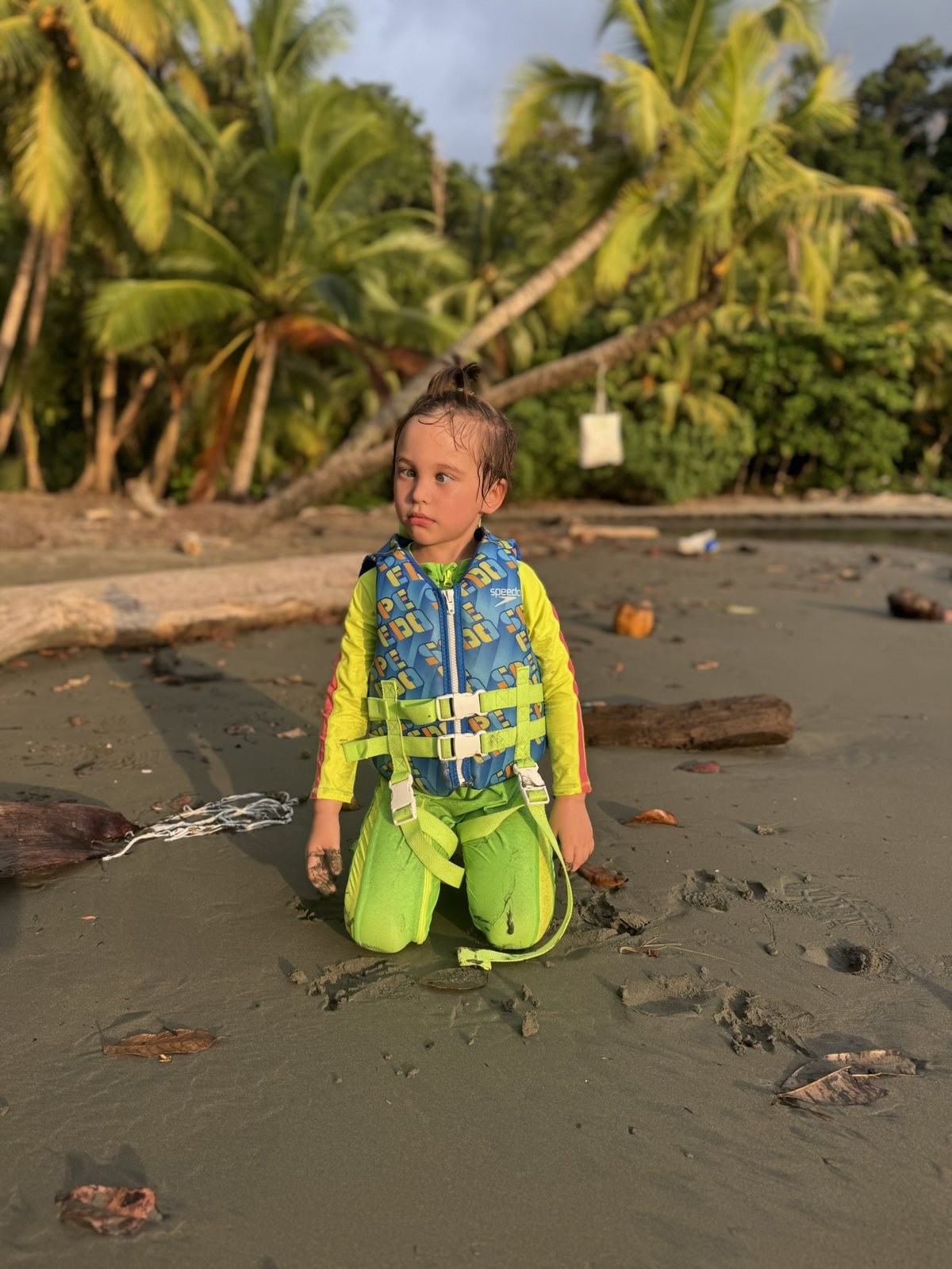 A five-year-old in a neon swimsuit and a lifejacket kneeling on the black sand at Playa Ventanas in March 2025, with driftwood and palms behind him