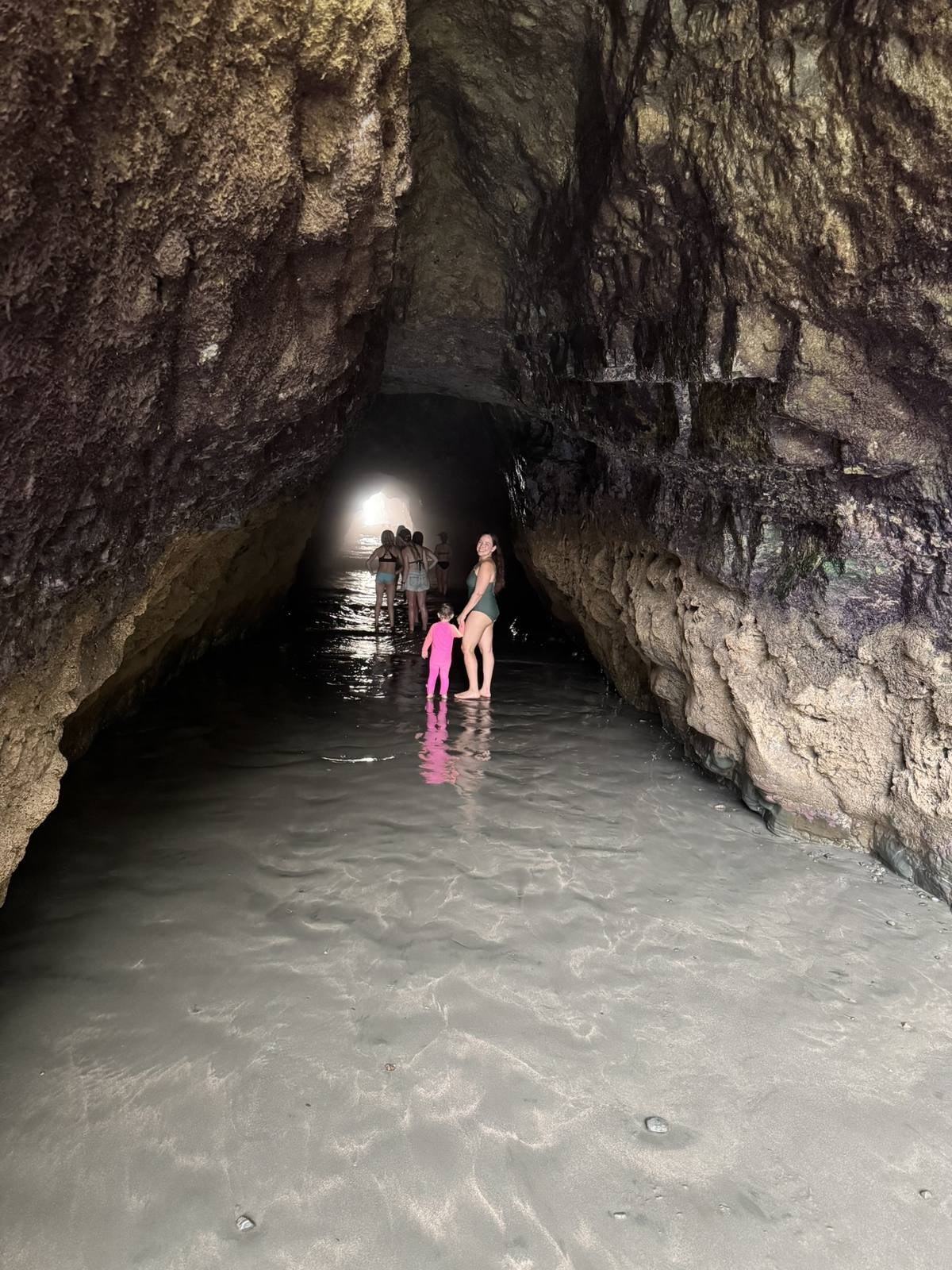 A mother leading her three-year-old daughter, in a neon pink swimsuit, through ankle-deep water inside the sea cave at Playa Ventanas in July 2025, with daylight showing at the far end of the tunnel