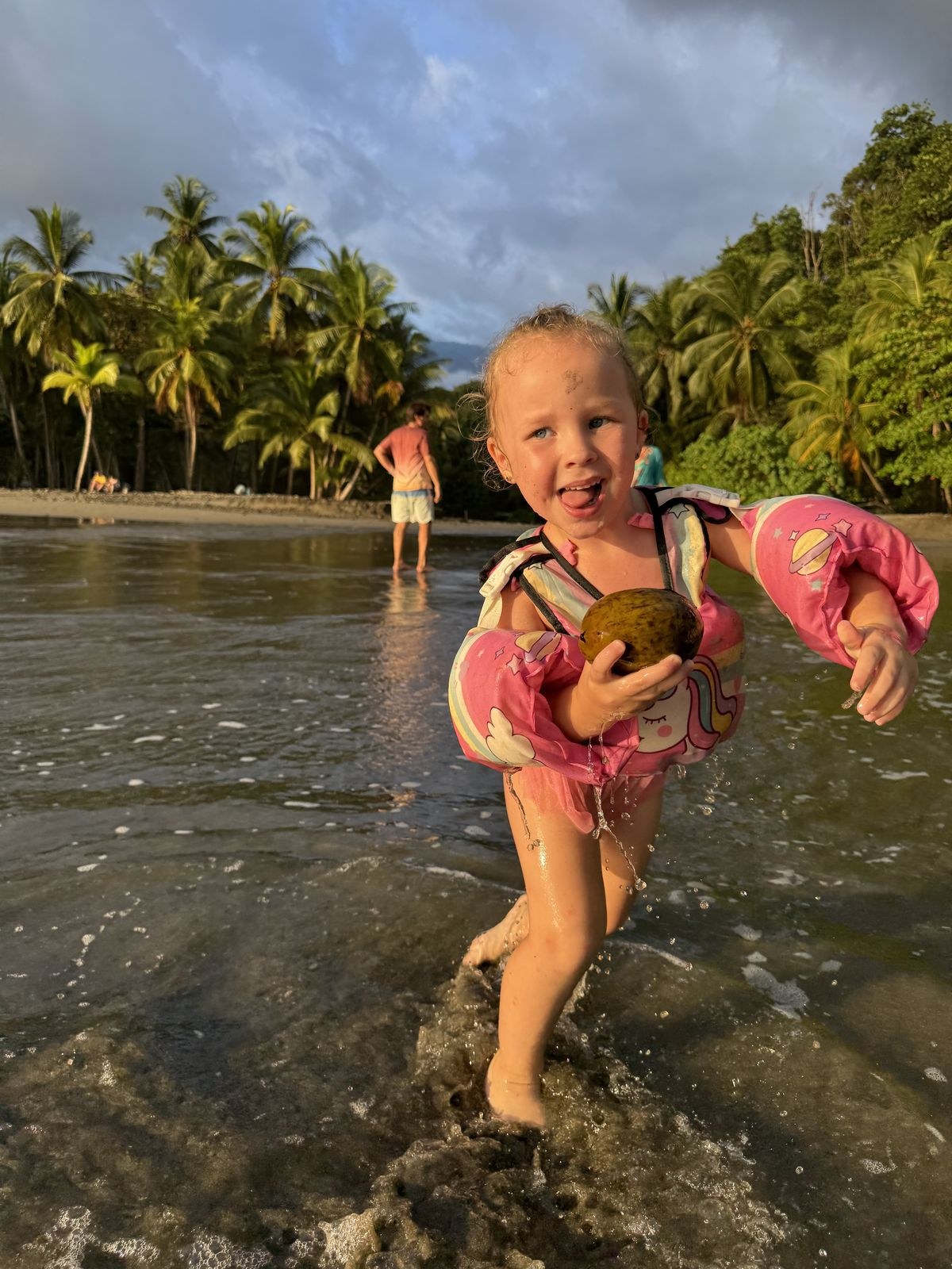 A four-year-old girl in a pink float vest carrying a coconut through the shallow water at Playa Ventanas, Costa Rica