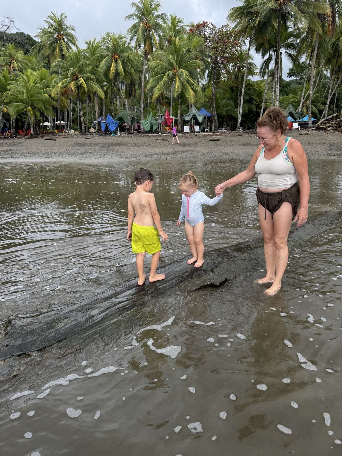 Lela holding her granddaughter's hand as both children walk along a driftwood log in the shallow river pool at Playa Ventanas, Ojochal, Costa Rica, with vendor tents and palms behind them