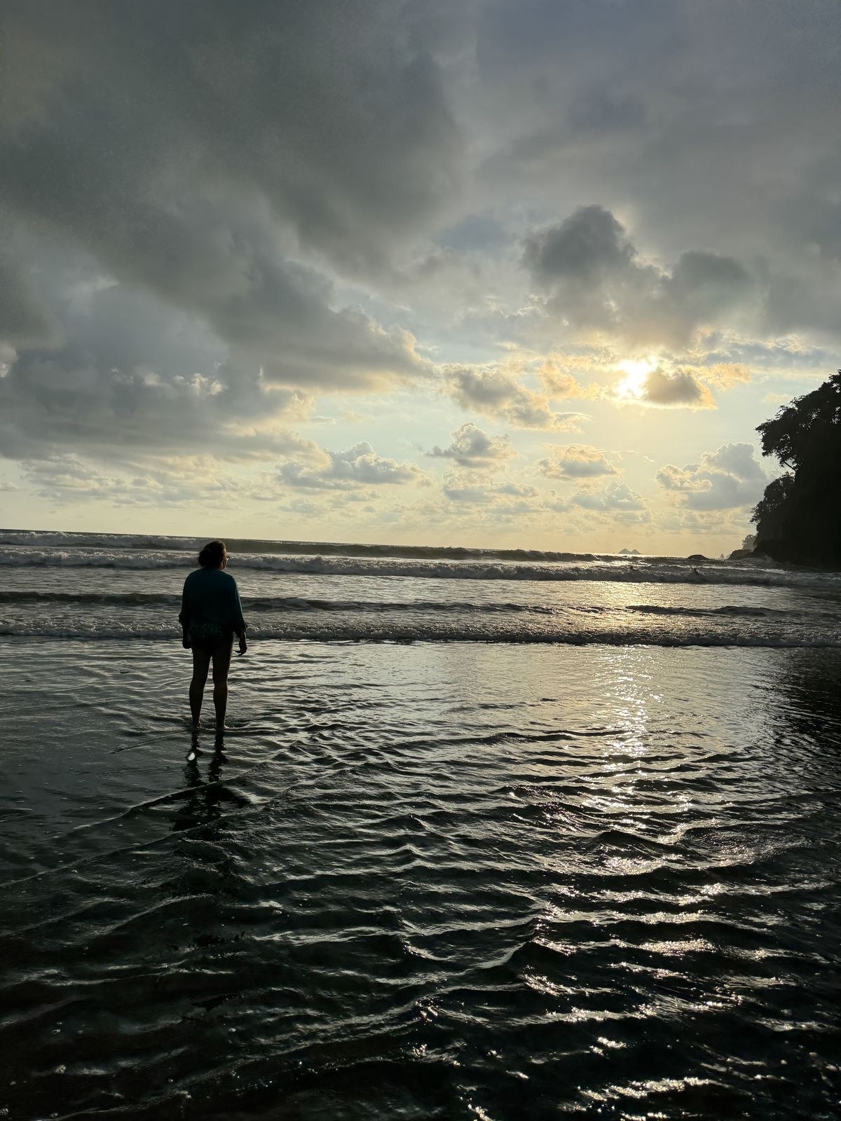 Lela standing in the shallow water at Playa Ventanas, Costa Rica, watching the sun go down over the ocean with the headland dark on the right