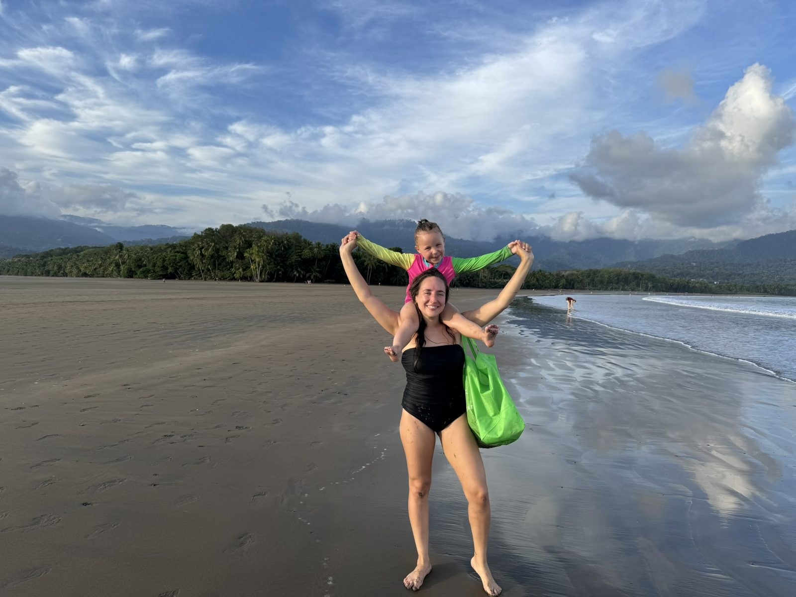 A mother standing on the wet sand with a four-year-old in a bright pink and green long-sleeved swim guard on her shoulders, forested mountains behind
