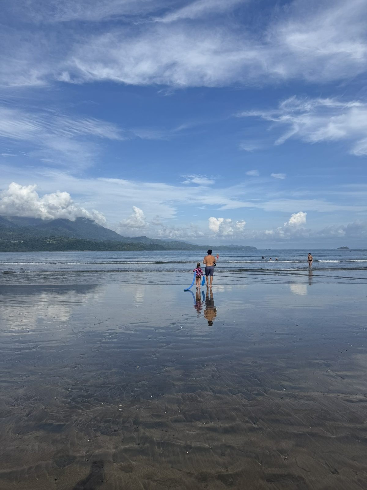 A man and a small child in a pink swim guard walking toward the water across a vast sheet of wet sand at low tide, mountains along the coast behind them