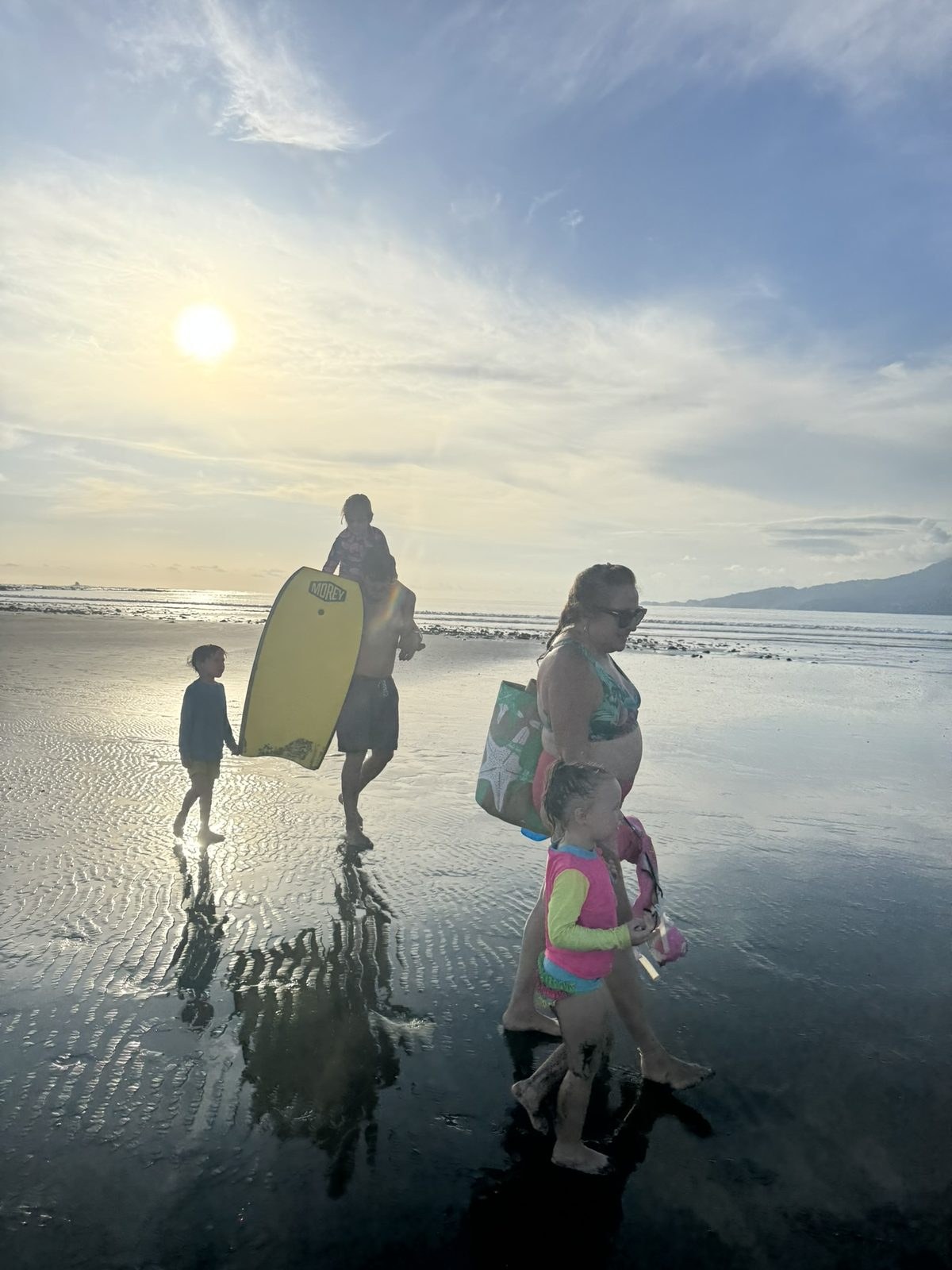 A family walking back across the wet low-tide flats carrying a yellow boogie board and a beach bag, one child riding on an adult's shoulders, low sun behind